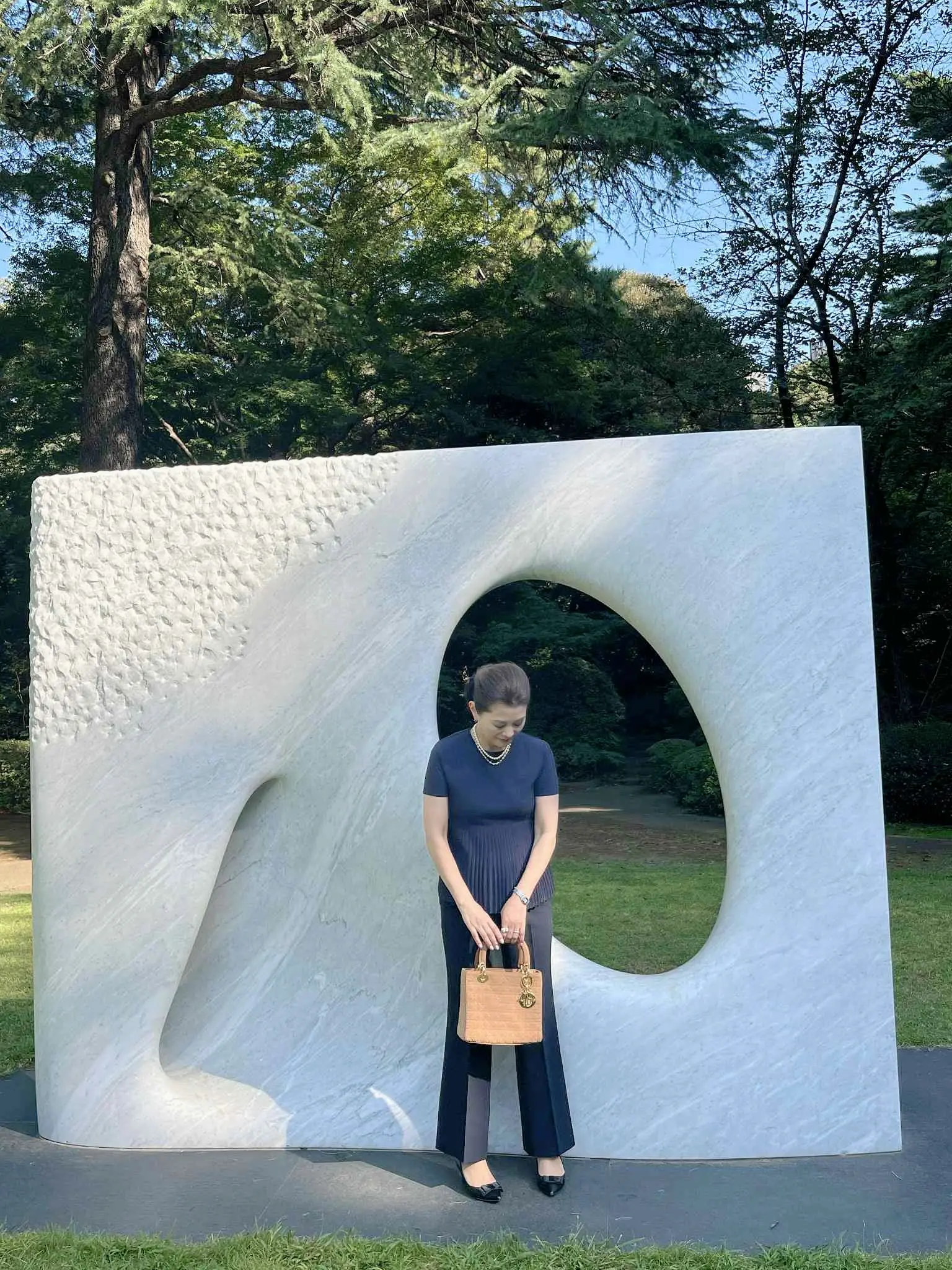 安田侃の大理石彫刻《風》の前でうつむきながら立つ女性。ネイビーの上品な装い。