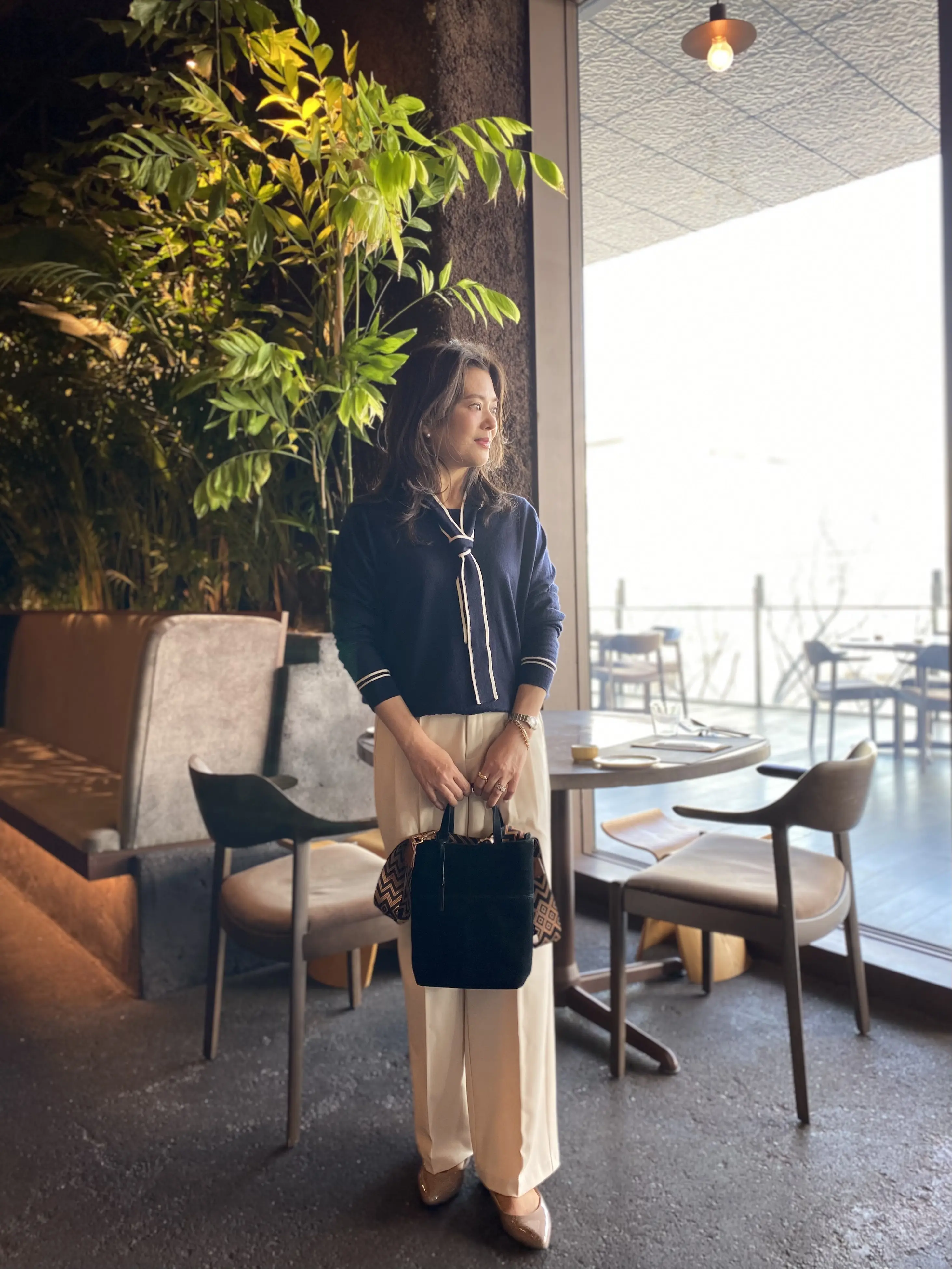 表参道ランチにおすすめのフレンチレストラン「bonélan（ボネラン）」。GYRE4階の緑に包まれた空間で、50代大人女性がジョンスメドレーのボウタイニットとGUタックワイドパンツ（白）を合わせたきれいめランチコーデを楽しむ様子