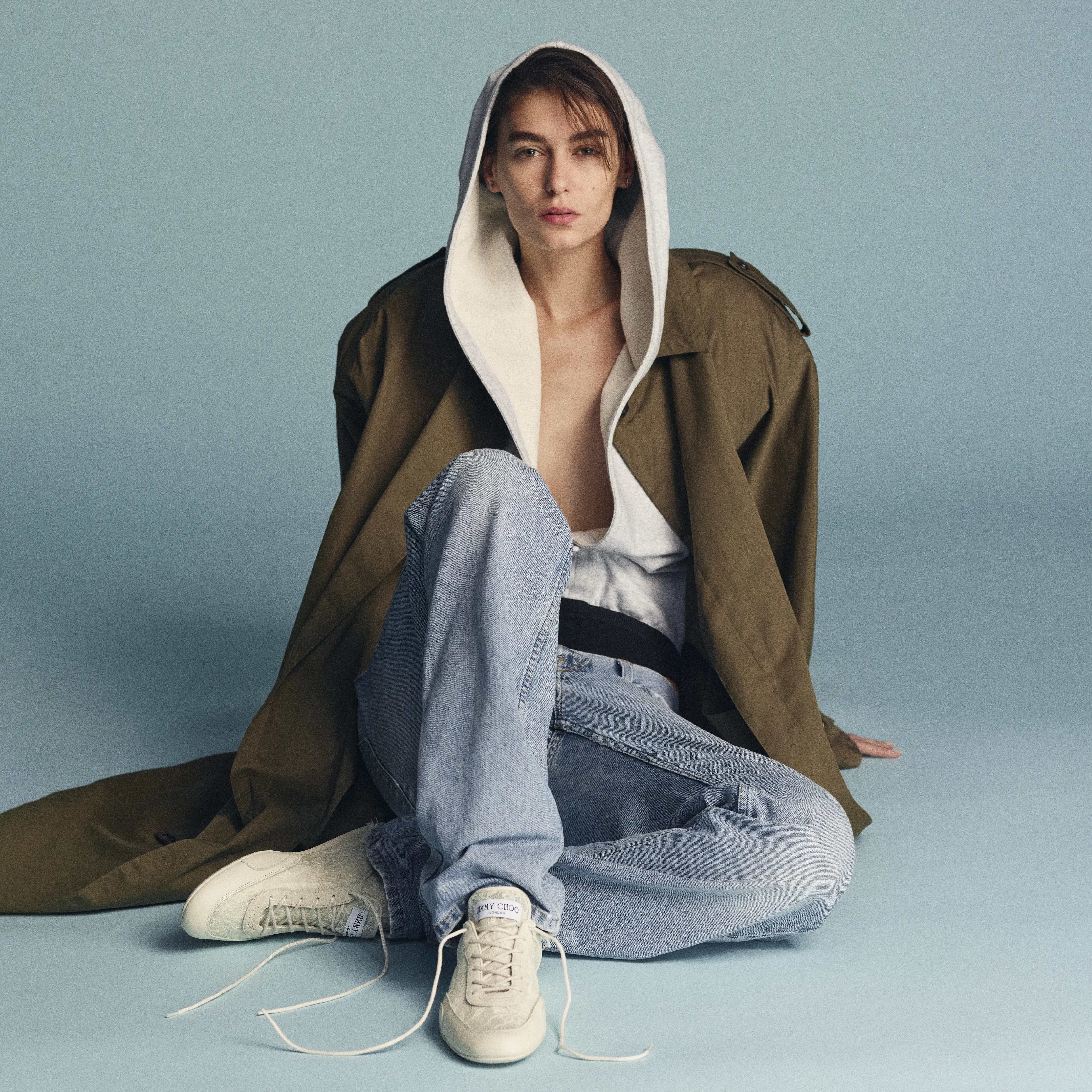 白レーススニーカー ジミー チュウ「SUNNY」を履いた女性モデルの写真。モデルはライトブルーのデニムパンツにグレーのパーカーを合わせ、その上からカーキのトレンチコートをはおったコーディネート