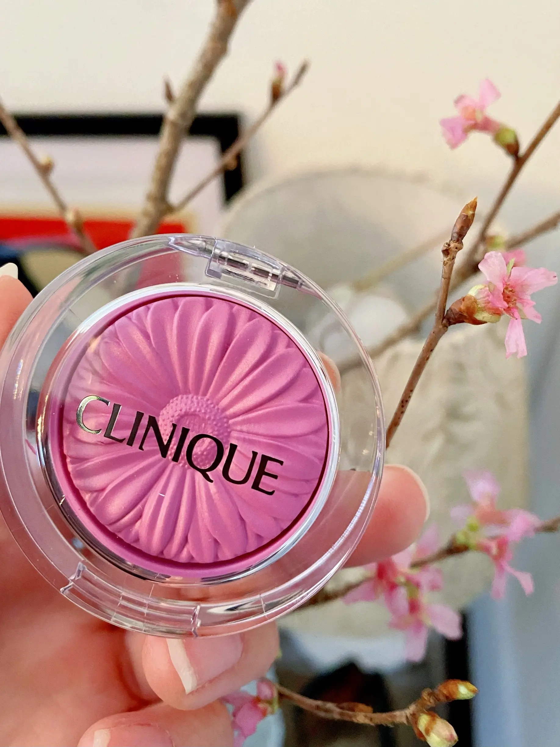 クリニーク　チークポップ 15　パンジーポップ CLINIQUE CHEEK POP  PANSY POP15を持つ女性の手と花の咲いた桜の枝の対比画像