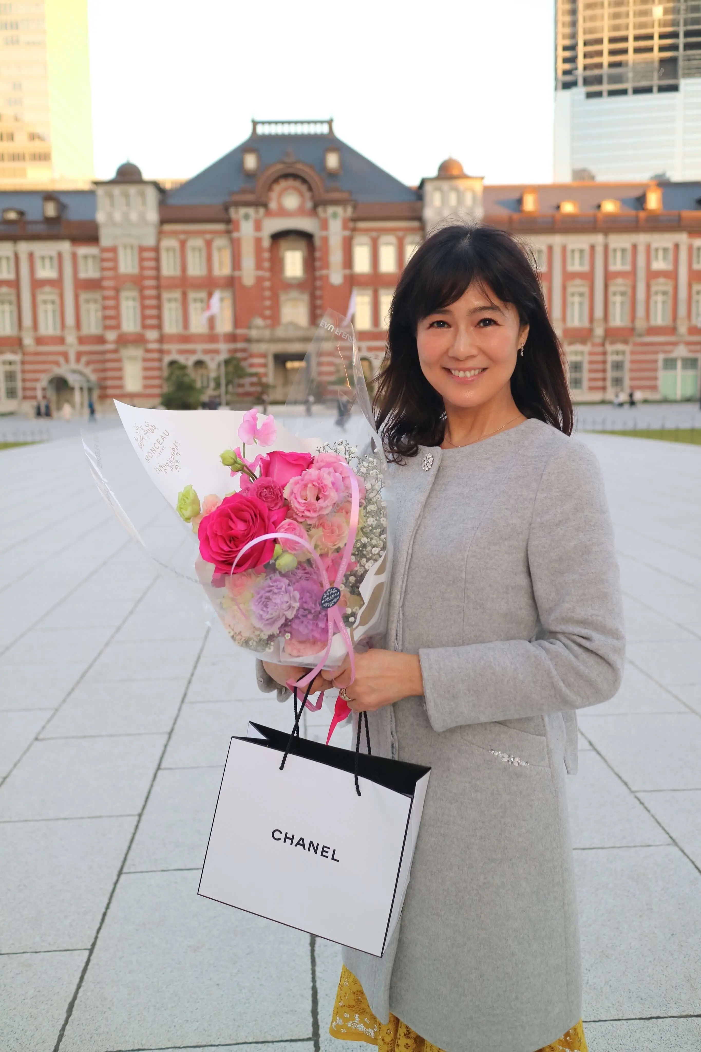 グレーのコートでお誕生日の花束とプレゼントを持って東京駅の前で記念撮影
