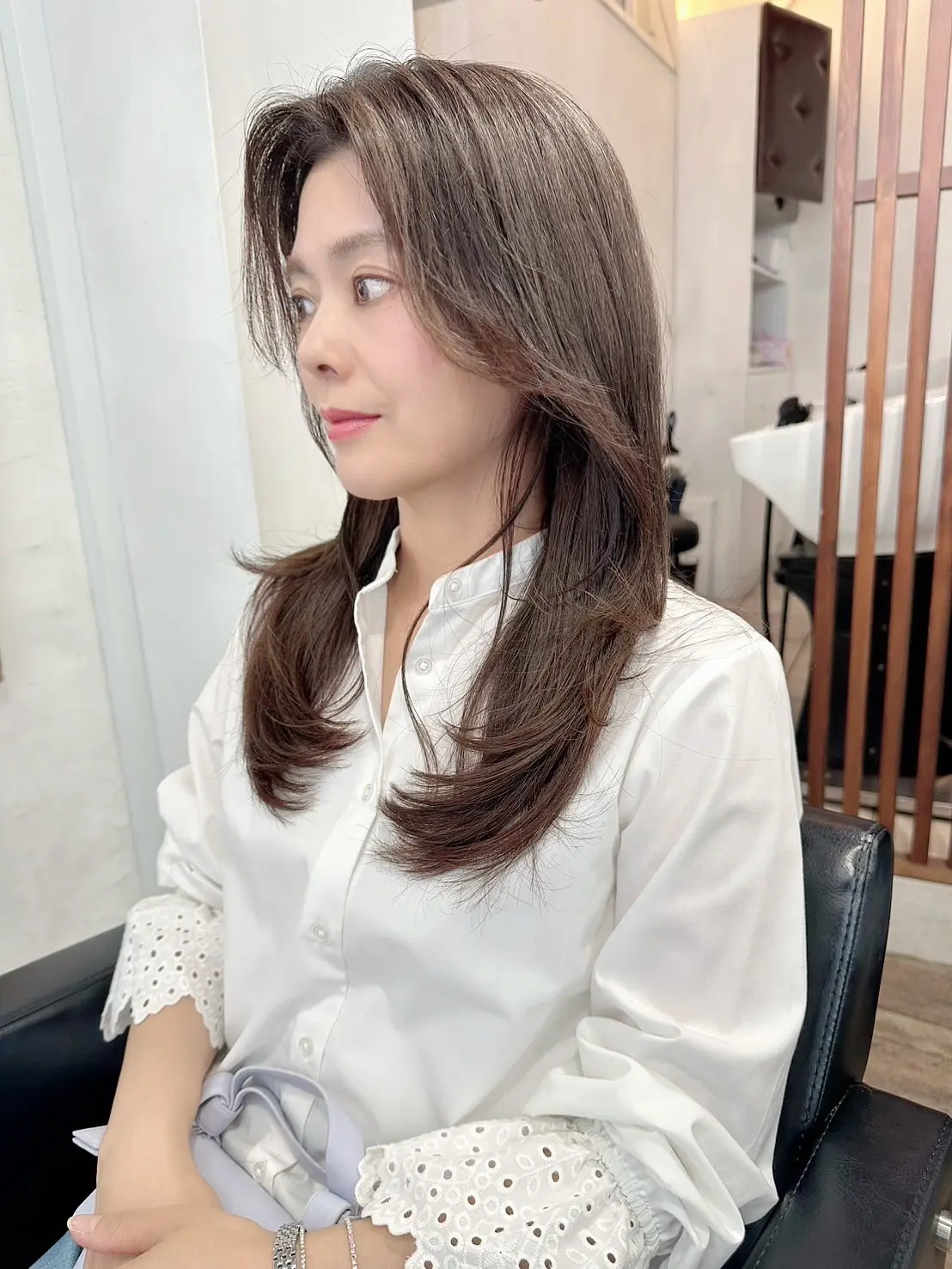 原宿「ハーツ」店内の椅子に座り、レイヤーが美しく入った柔らかなロングヘアで白いブラウスに身を包んだ女性が、自然光の中で横顔を見せている。