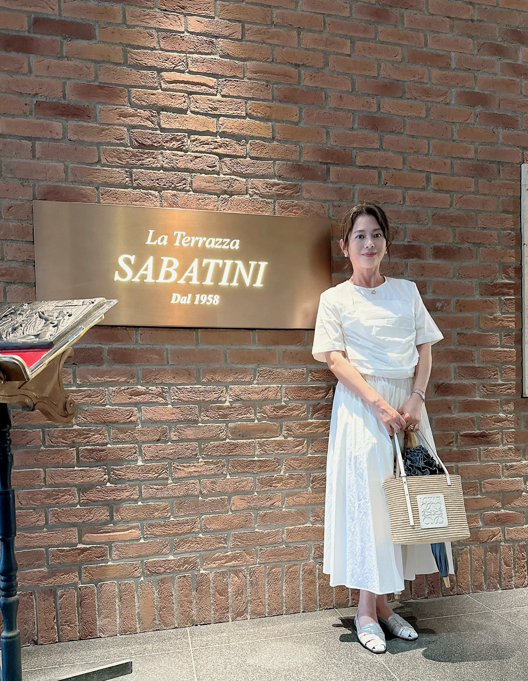 白のワンピースとロエベのかごバッグでまとめた女性の夏コーデ。La Terrazza SABATINIの前で撮影された。