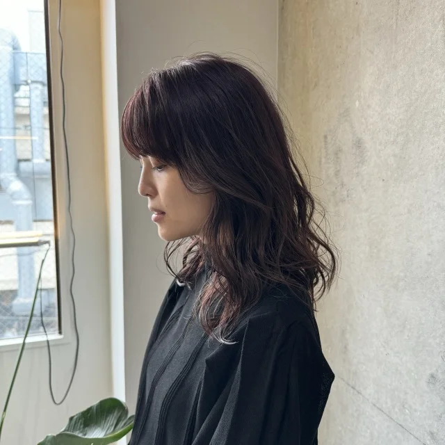 Side profile of a woman with wavy, shoulder-length hair wearing a black top, standing near a window with a potted plant in the foreground