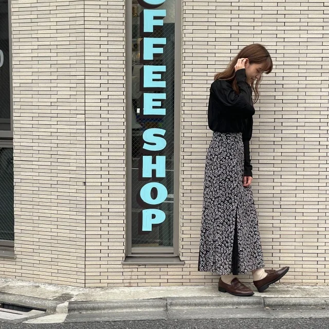 “Full-body shot of a woman standing in front of a beige brick wall with a vertical ‘COFFEE SHOP’ sign in blue. She is wearing a black top, a long patterned skirt, and brown loafers, looking down while touching her hair.”