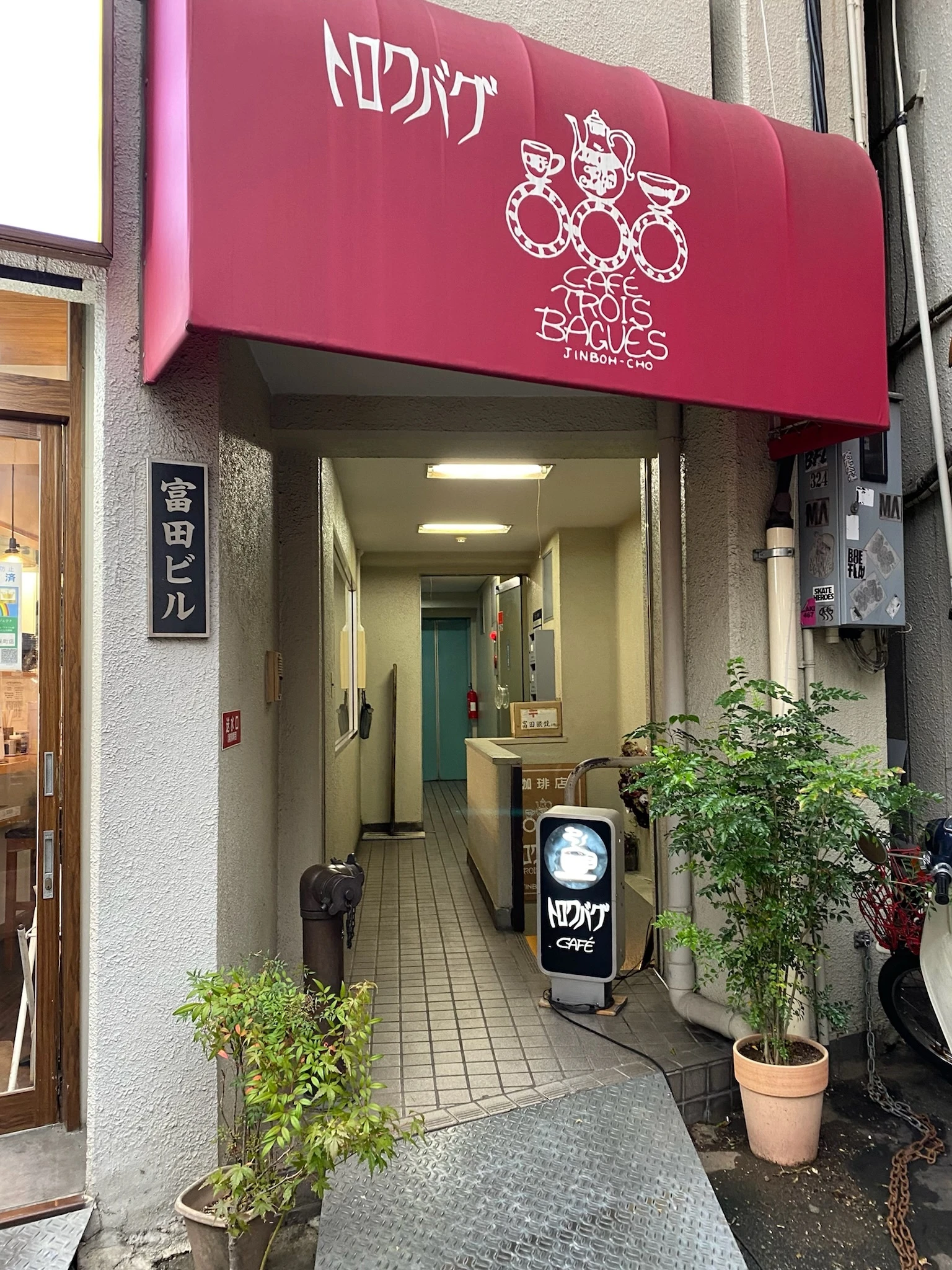東京都神保町駅の近くにある喫茶店「トロワバク」のお店の外観の写真です。地下へ伸びる階段の前に植物と小さな看板が置かれています。看板には店名とコーヒーのイラストが描かれています。