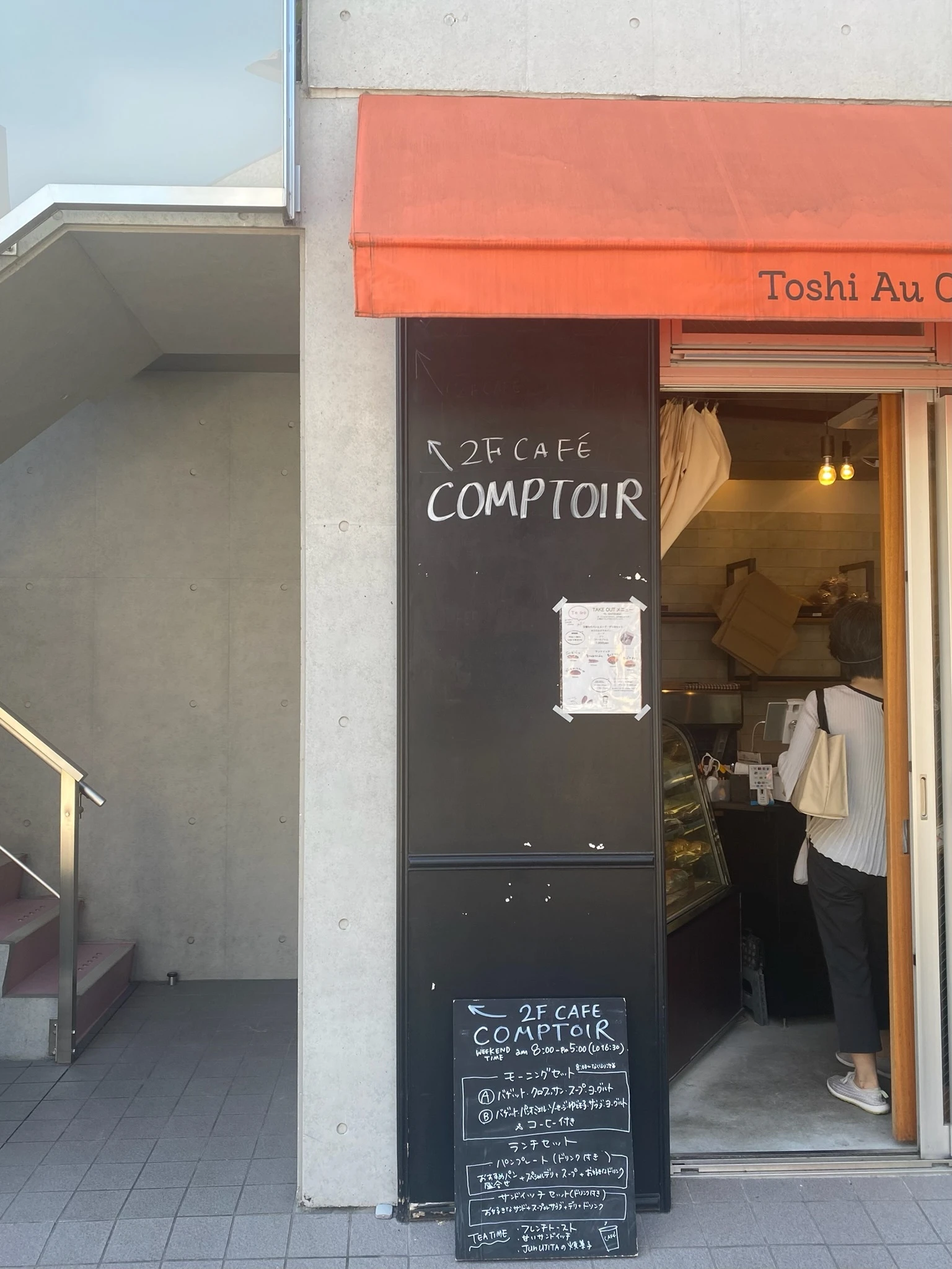 都立大学駅にあるパン屋さん「Toshi Au Coeur du Pain」の店構え。外観。オレンジ色のひさしには、黒で「Toshi Au Coeur du Pain」の文字がプリントされています。入り口横の黒板には、白のチョークで2階にある「COMPTOIR」への案内が描かれています。