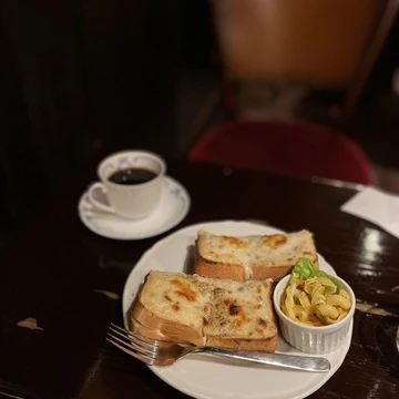 本のまちで知られる東京都神保町にある喫茶店「トロワバグ」。創業以来の人気メニュー「グラタントースト」とコーヒーのセットの写真。暗い店内。濃い茶色のテーブルの上にグラタントーストと、マカロニサラダが乗った白いお皿があります。左奥には、白いマグカップに入ったコーヒーが置かれています。
